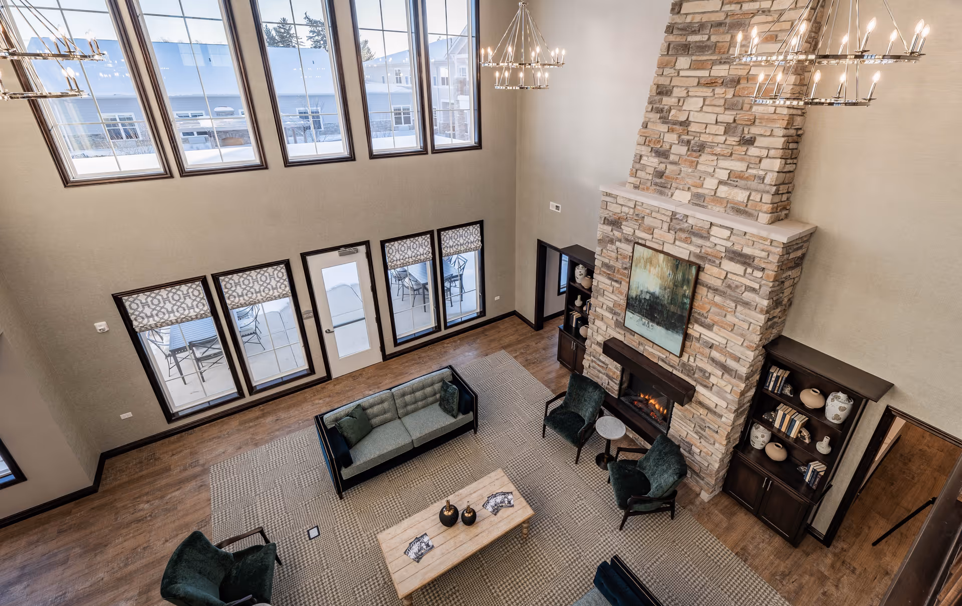 A spacious senior living facility common area with high ceilings, large windows, and a stone fireplace. The room features a green sofa, two green armchairs, a wooden coffee table, and bookshelves with decorative items. Outside, a patio with tables and chairs is visible through the windows.