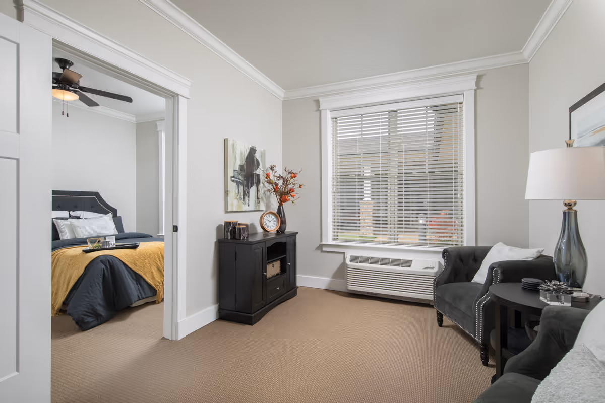 A cozy living room area with a dark armchair, a side table with a lamp, and a black cabinet adorned with a clock, vase with flowers, and decorative items. A large window with white blinds lets in natural light. Through an open doorway, a bedroom is visible with a bed covered in dark bedding and a mustard yellow throw, a ceiling fan, and neutral-colored walls.