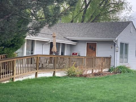 Single-story white building with a gray shingled roof, a wooden ramp with railings leading to a brown door, surrounded by green grass and trees in the background.
