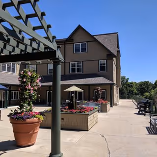 Outdoor patio with a pergola, potted and raised flower planters in front of a three-story beige building under a clear blue sky.
