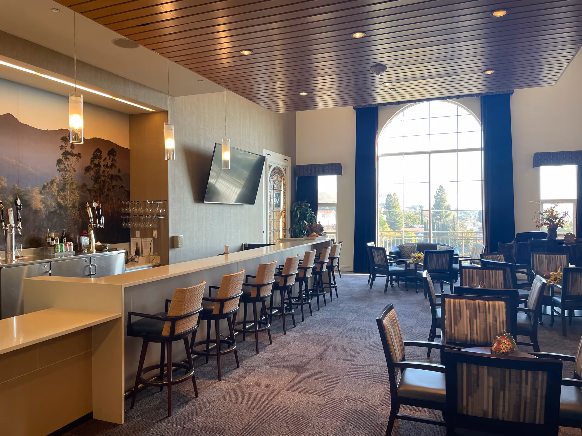 A spacious and well-lit lounge area with a long bar counter and several bar stools on the left side. Behind the bar are beer taps and a large mural of a mountain landscape. To the right, there are multiple tables and chairs arranged for seating, with large windows letting in natural light and offering a view of trees outside. The ceiling has wooden paneling with recessed lighting.