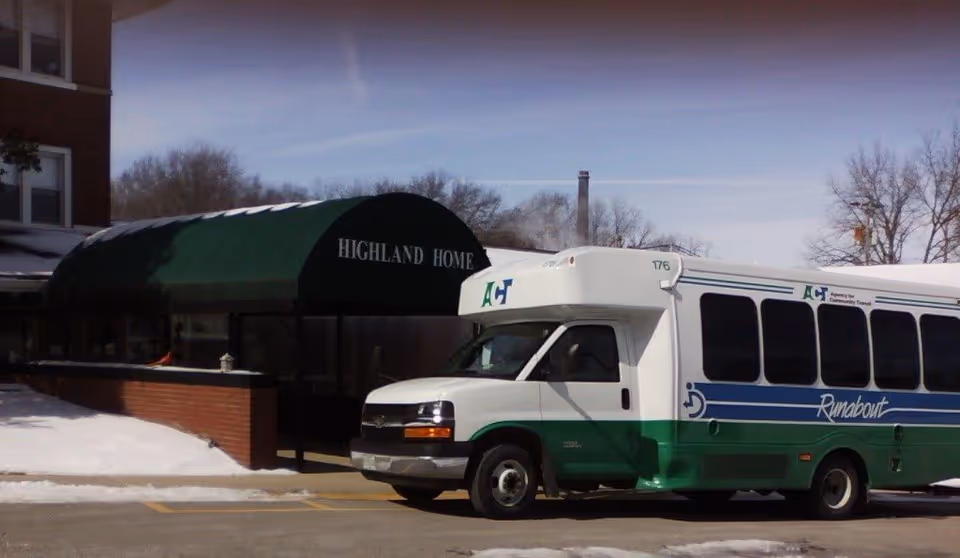 A white and green shuttle bus labeled 'Runabout' parked outside the entrance of a building with a green awning that reads 'Highland Home'. Snow is visible on the ground and leafless trees are in the background.