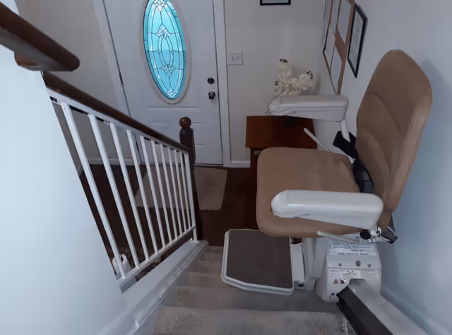 A stairway with a beige stair lift chair installed on the right side. At the bottom of the stairs is a white door with an oval-shaped decorative glass window. The stairway has a white railing on the left side and a small brown table with decorative items near the door.