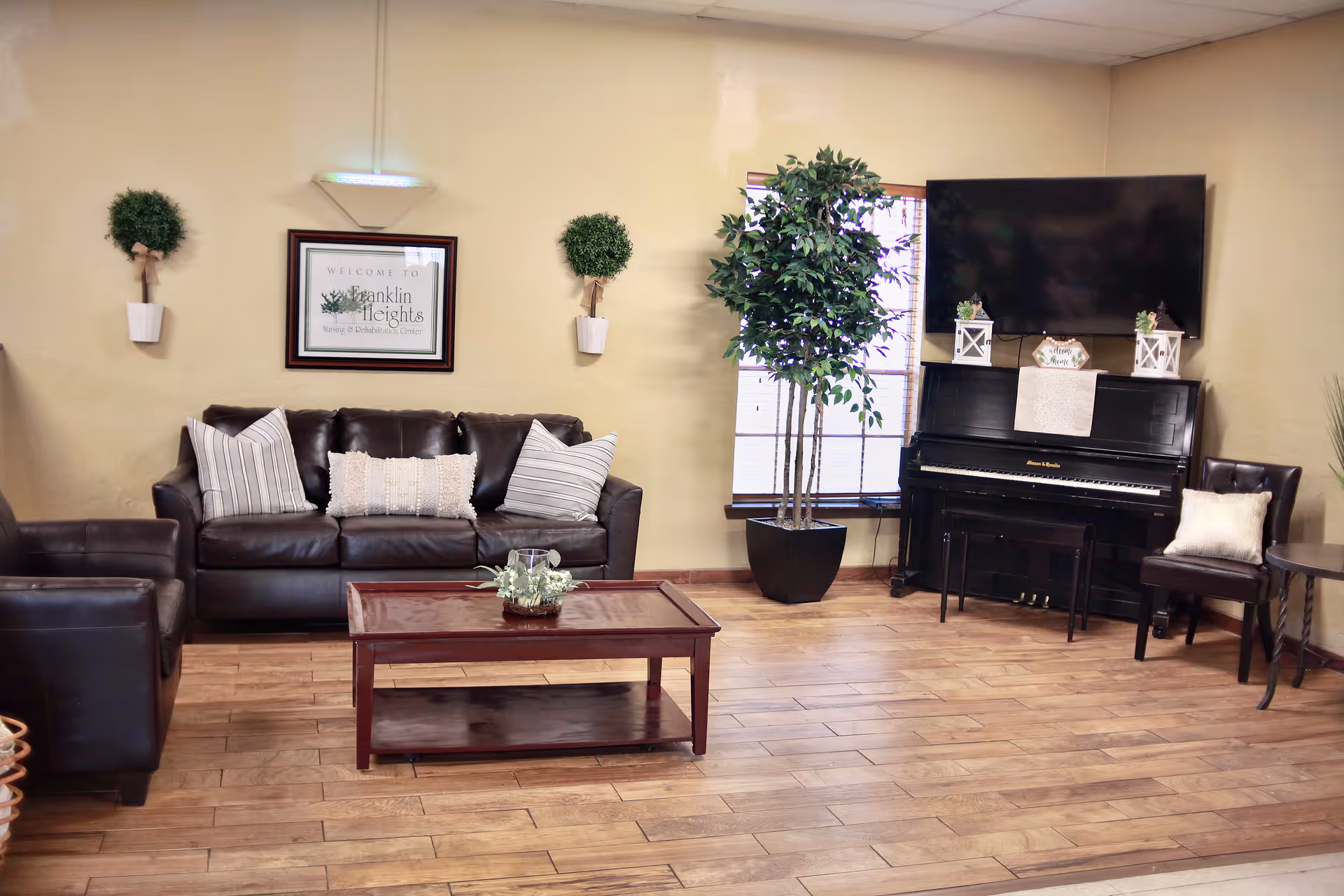 A cozy living room area with a dark brown leather sofa and armchair, a wooden coffee table with a small plant centerpiece, two wall-mounted potted plants, a large potted tree near a window, a black upright piano with decorative items on top, a flat-screen TV mounted above the piano, and a dark brown chair with a light-colored pillow next to a small round table.