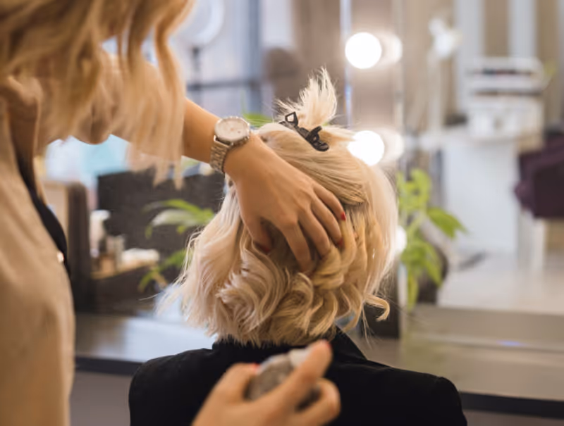 A person styling the blonde hair of a seated woman in a salon setting, with a mirror and lights in the background.