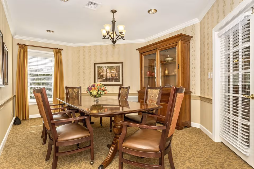 Formal dining room with a wooden table and chairs, a china cabinet, chandelier, window with gold curtains, and a floral centerpiece.