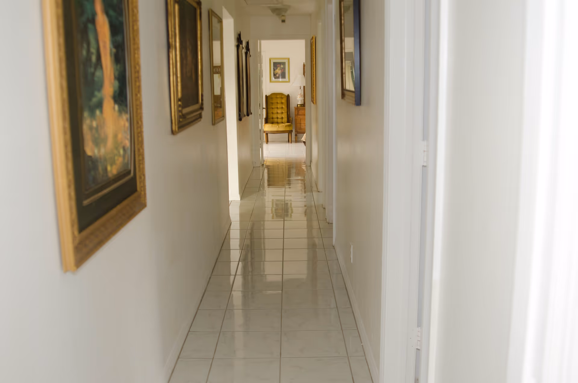 A long, narrow hallway with white tiled floor and white walls adorned with framed paintings. At the end of the hallway, there is a yellow upholstered chair next to a wooden side table with a lamp and a framed picture on the wall.