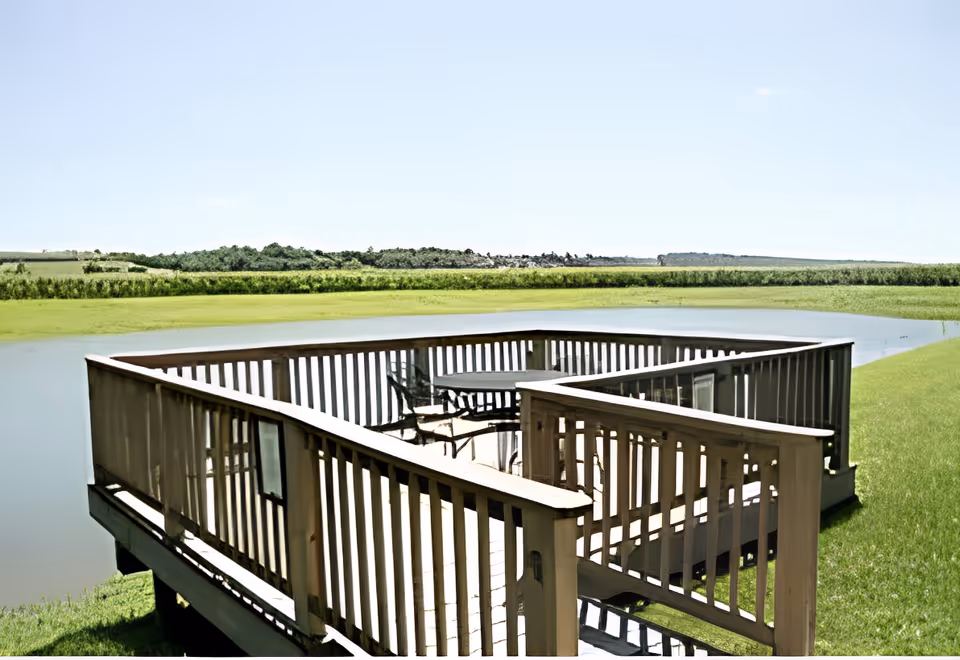 A wooden deck with railing overlooking a calm pond surrounded by green grass and distant trees under a clear blue sky. The deck has a round table and several chairs.
