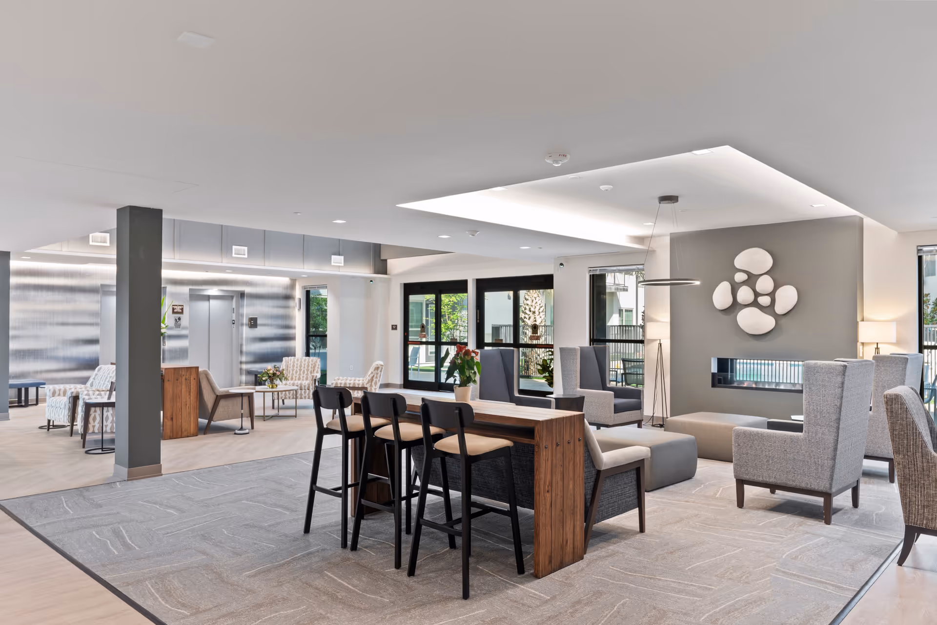 A modern and spacious senior living facility common area with a mix of seating options including high chairs at a wooden counter, armchairs, and cushioned benches. The room features large windows letting in natural light, a contemporary wall-mounted fireplace, and decorative wall art above the fireplace. The flooring is a combination of carpet and wood, and there is an elevator visible in the background.