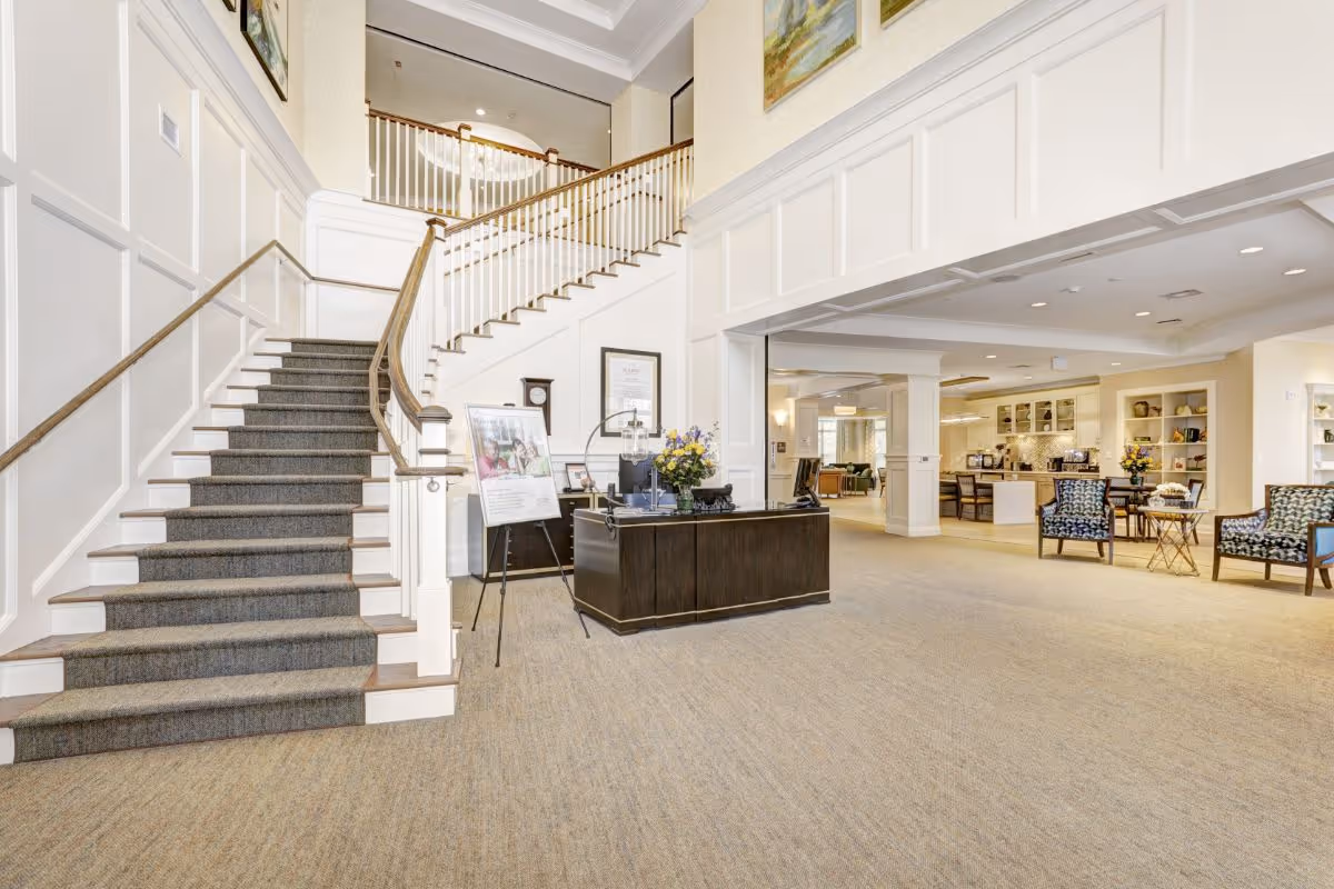 Bright and spacious interior of a senior living facility featuring a carpeted staircase with wooden handrails, a dark wooden reception desk with flowers and informational materials, and a seating area with patterned chairs and tables. The area is well-lit with recessed lighting and has white paneled walls and beige carpeting.