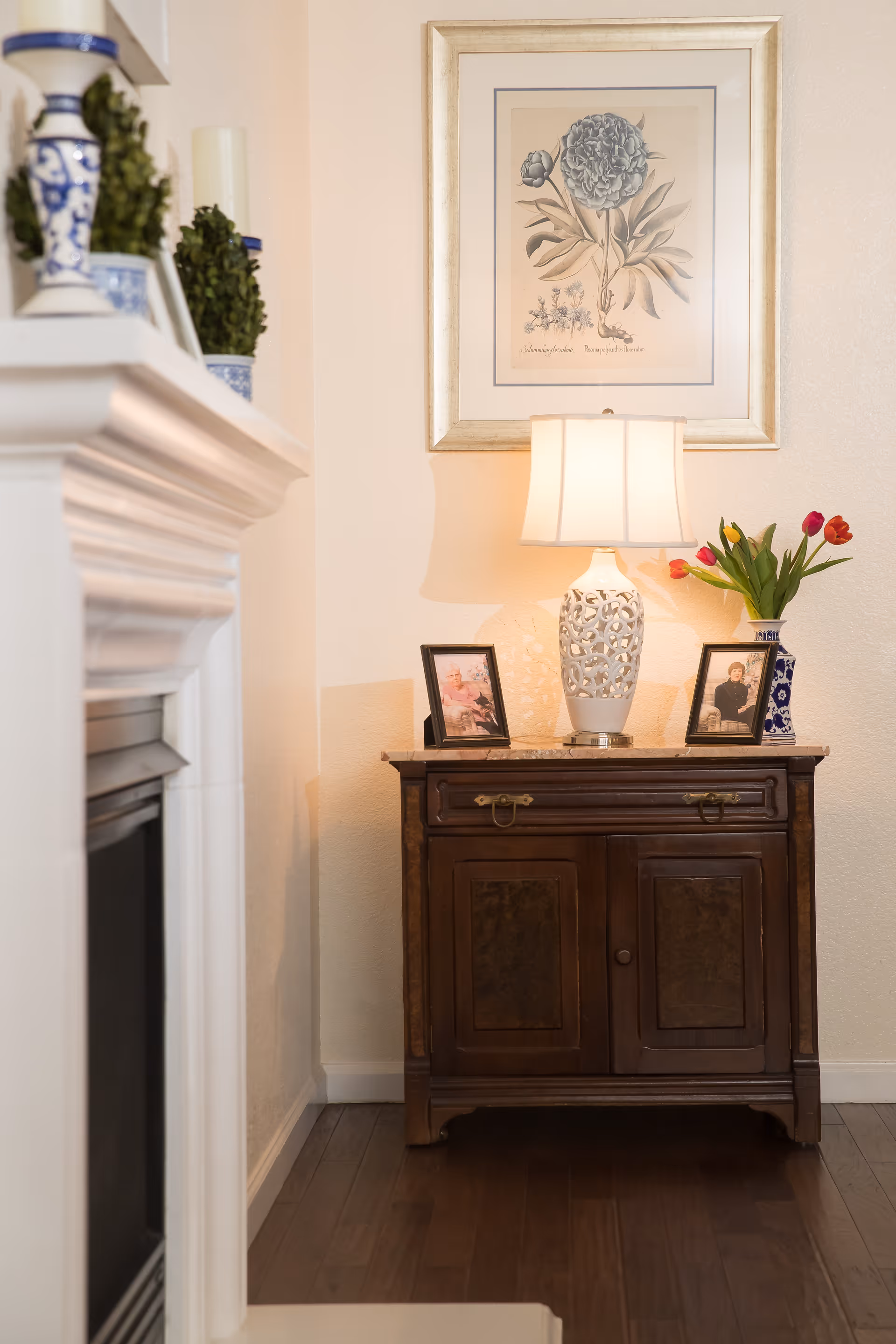 A cozy corner of a living room featuring a wooden cabinet with two framed photos, a decorative white table lamp, and a vase with red tulips. Above the cabinet hangs a framed botanical print. To the left, part of a white fireplace mantel with blue and white decorative items and small green plants is visible. The floor is dark wood.