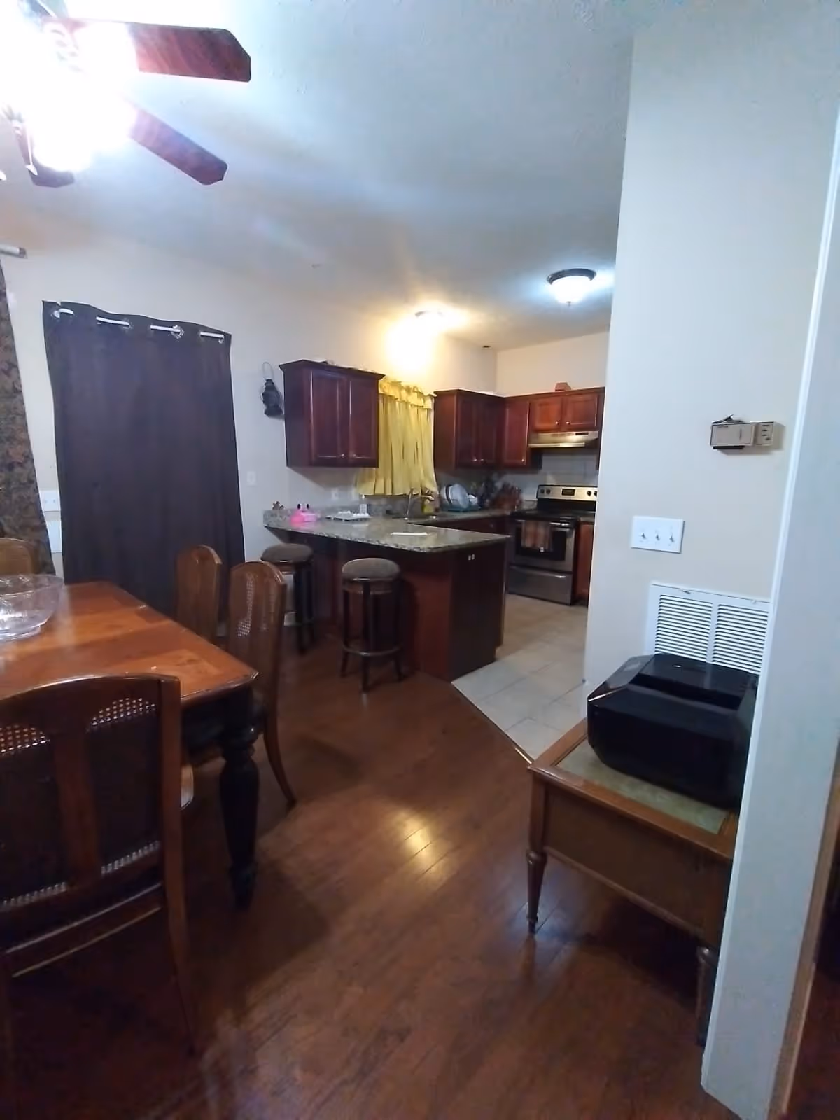 Interior view of a home showing a dining area with a wooden table and chairs on the left, and a kitchen area with wooden cabinets, a granite countertop island with stools, and stainless steel appliances in the background. There is a ceiling fan with lights above and a small table with a black appliance on the right side.