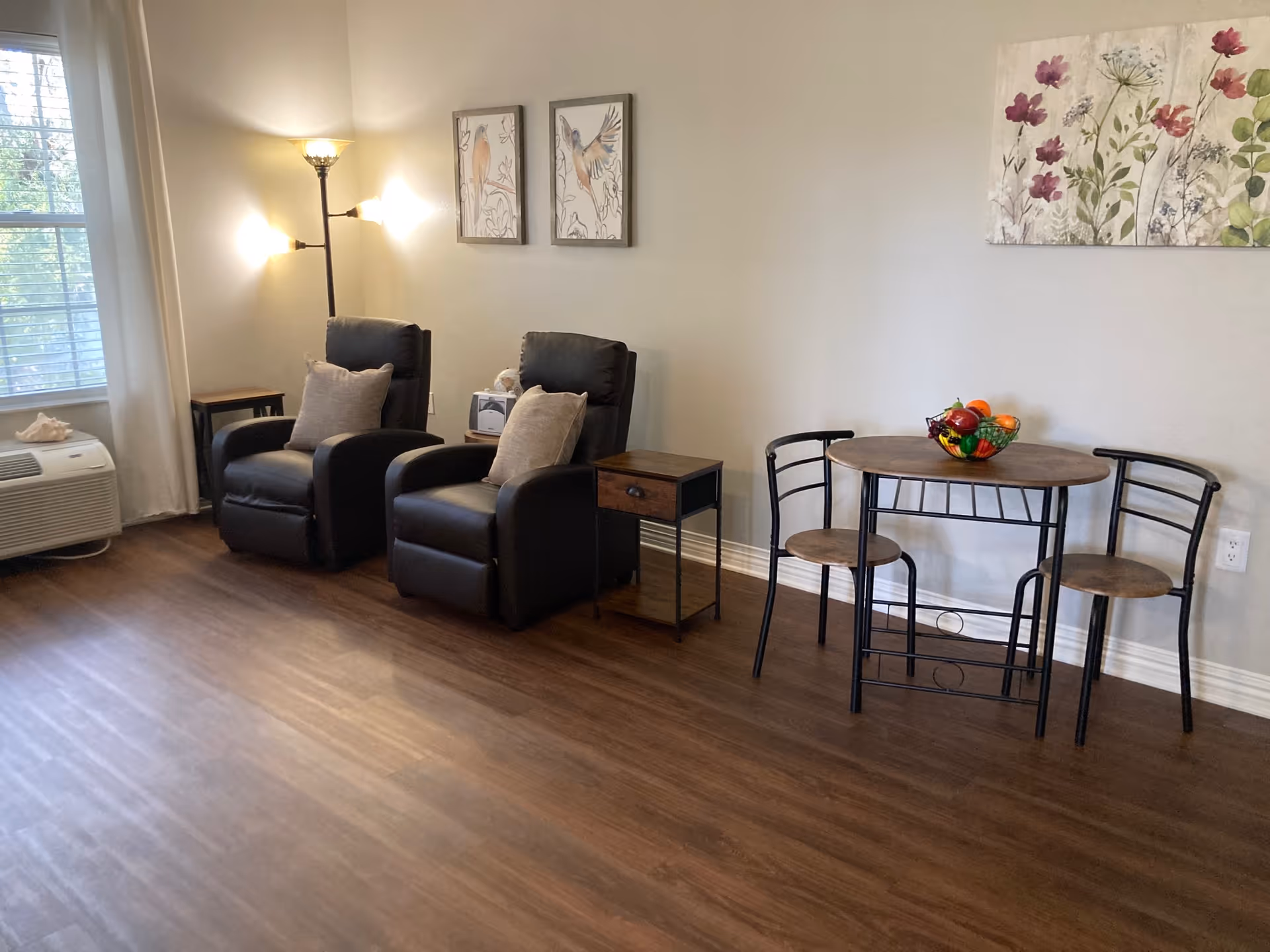 A cozy interior room with two black recliner chairs each with a beige pillow, a small wooden side table between them, a tall floor lamp with three lights, and a small round wooden table with two matching chairs. The walls are decorated with framed bird artwork and a floral painting. There is a window with white curtains and an air conditioning unit below it. The floor is wood laminate.