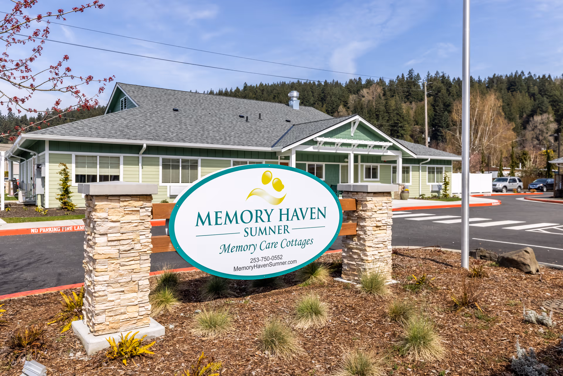 Exterior view of Memory Haven Sumner Memory Care Cottages building with a large sign in front displaying the facility name, phone number, and website. The building is light green with white trim, surrounded by a parking lot and landscaping with trees and bushes under a clear blue sky.