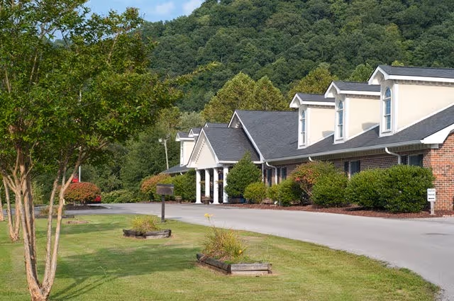 Exterior view of a single-story brick building with white trim and multiple dormer windows, surrounded by green bushes and trees, with a paved driveway and a grassy area with small planter boxes in front. A wooded hill is visible in the background under a clear sky.