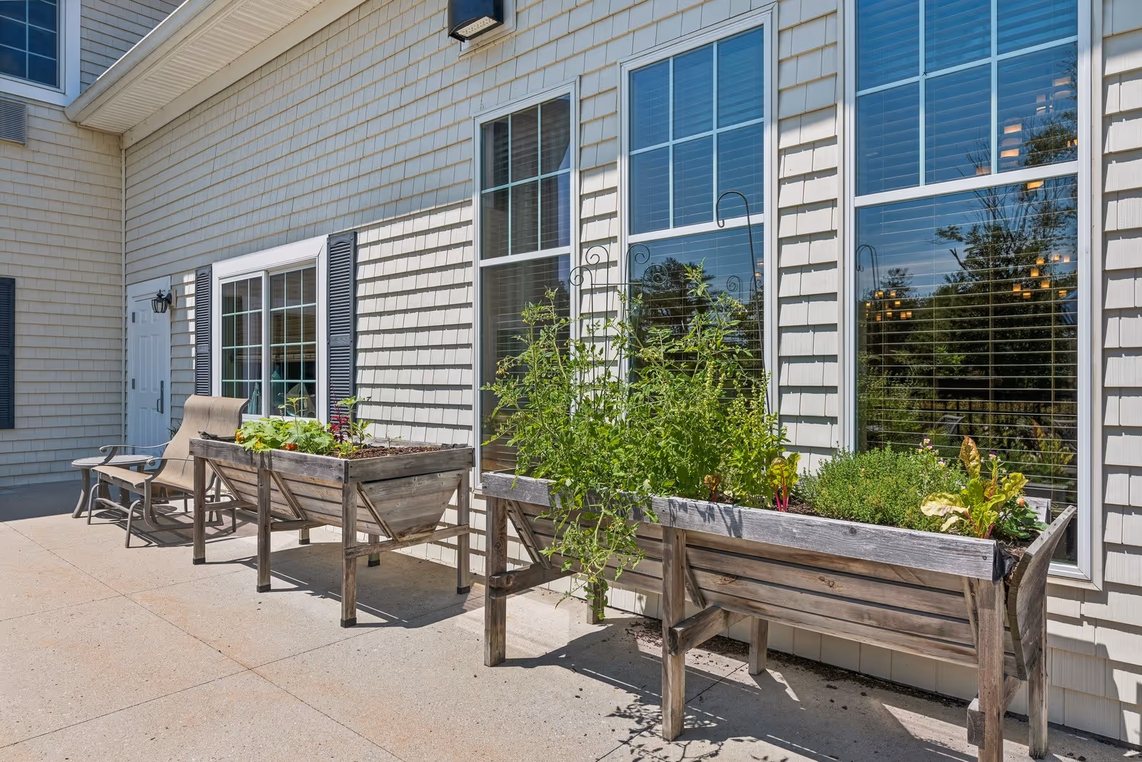 Outdoor patio area with two wooden raised garden beds containing various plants and herbs, positioned against the exterior wall of a building with large windows and white siding. There are two beige cushioned chairs and a small table nearby on the concrete patio.