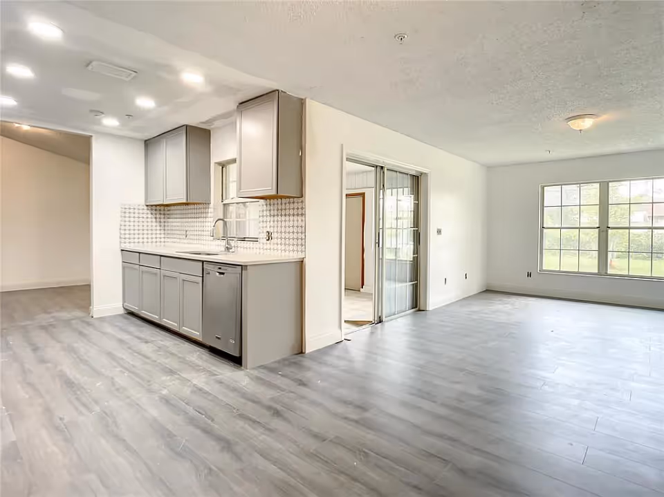 Bright open-plan interior showing a gray kitchen with cabinets and dishwasher adjacent to a spacious living area with large windows and wood-look flooring.