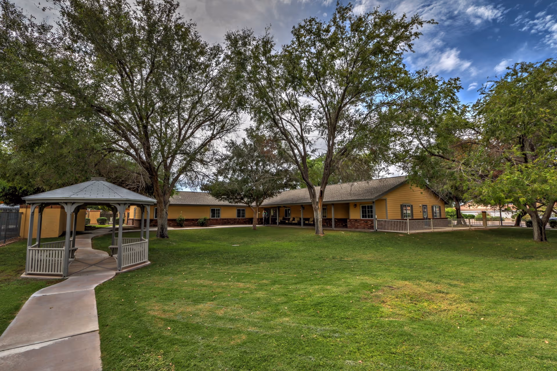 A spacious outdoor area with green grass, several large trees, a paved walkway leading to a small gazebo, and a single-story yellow building with a covered porch in the background under a partly cloudy sky.