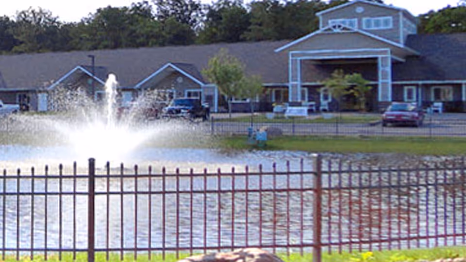 View of a senior living facility building behind a pond with a water fountain in the center, surrounded by a metal fence and some parked cars near the entrance.