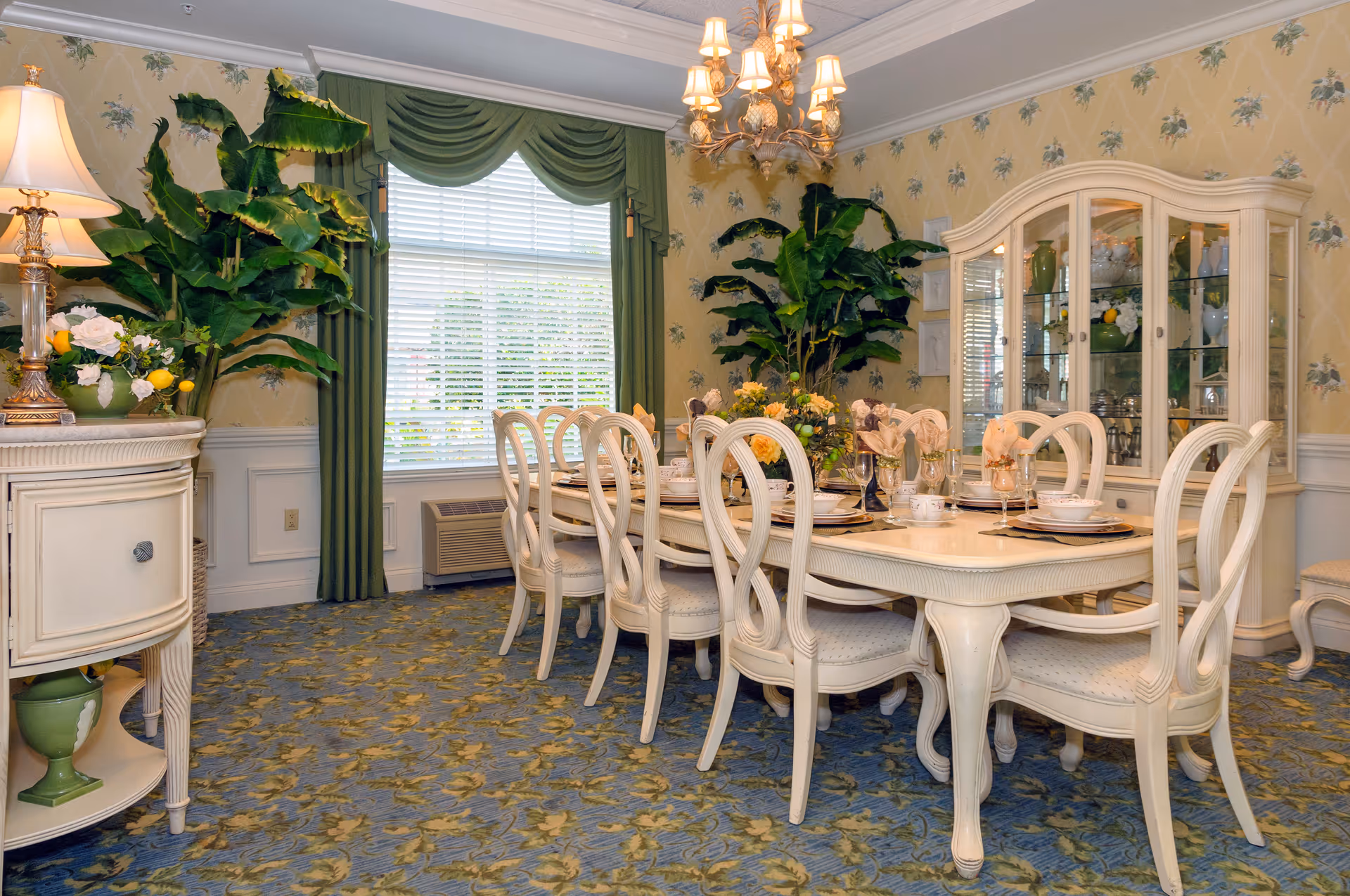 A formal dining room with a long white dining table surrounded by eight matching chairs. The table is set with plates, glasses, and napkins, and decorated with floral centerpieces. The room features floral wallpaper, a blue and yellow patterned carpet, green drapes on the window, and a chandelier hanging above the table. There are large green plants in the corners and a white china cabinet filled with dishes and glassware.