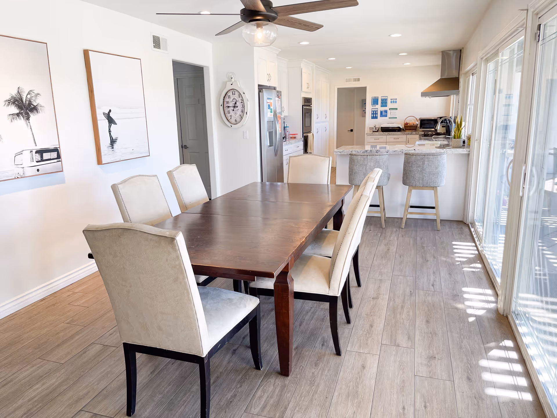 Bright dining area with a dark wooden table surrounded by six beige upholstered chairs. The room has light wood flooring and white walls adorned with two framed black and white pictures. In the background, there is a modern kitchen with white cabinets, stainless steel appliances, a wall clock, and a kitchen island with two gray bar stools. Large sliding glass doors on the right let in natural light.