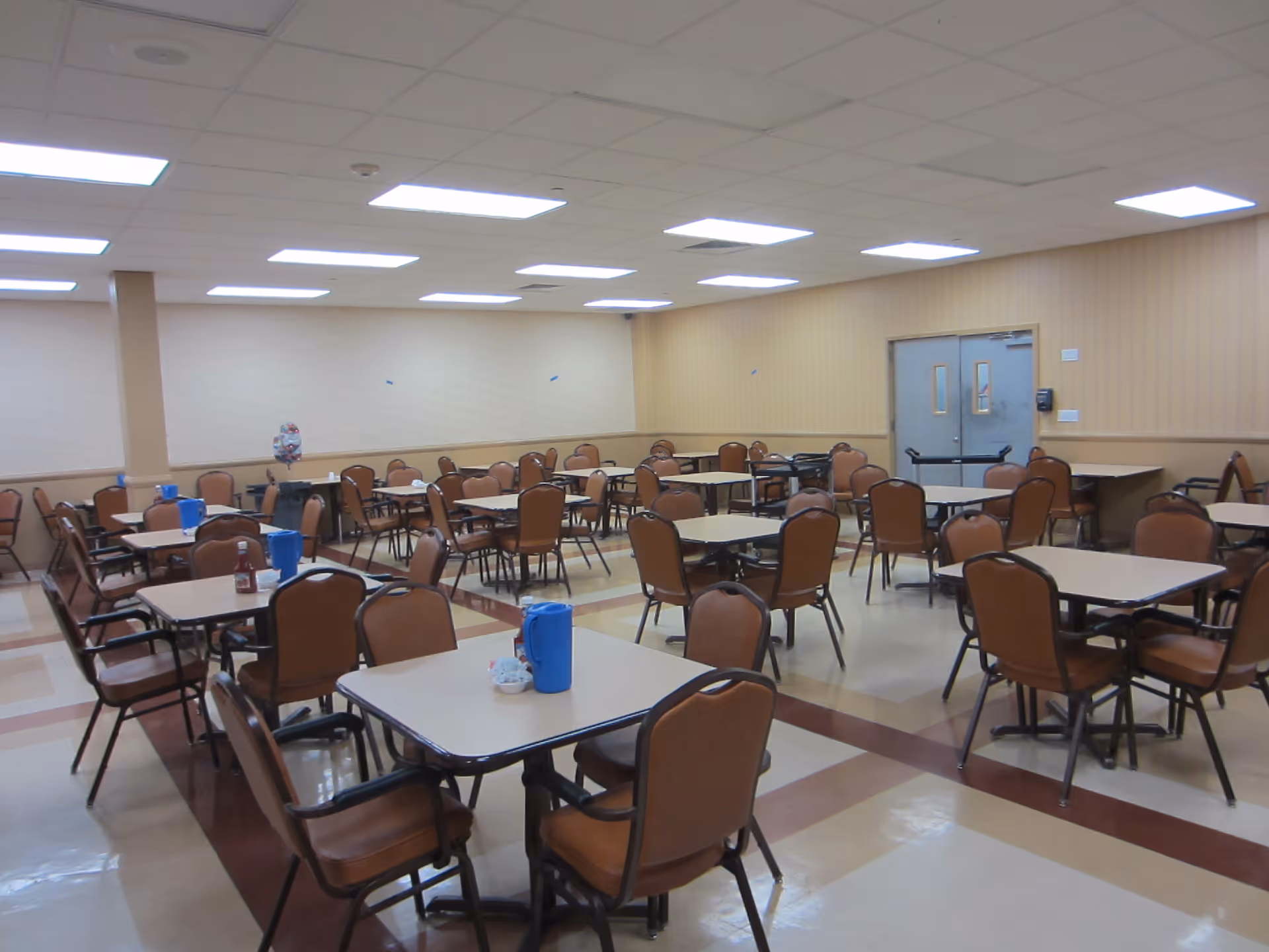 A large dining room with multiple square tables and brown cushioned chairs arranged neatly. The room has a tiled floor with a pattern, beige walls, and a drop ceiling with fluorescent lights. There are blue pitchers and condiment containers on some tables, and double doors are visible at the back of the room.