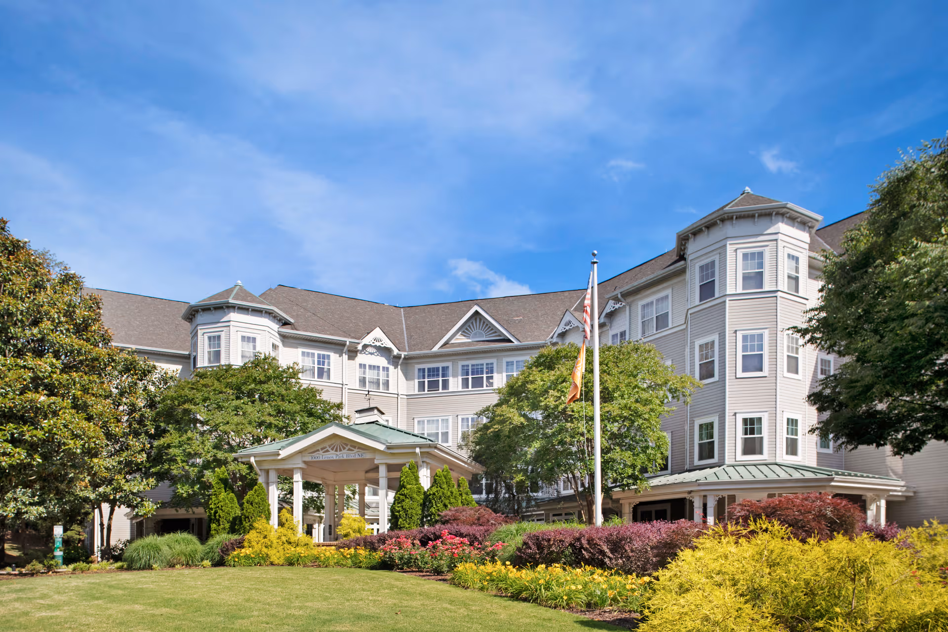 Exterior view of a large, multi-story senior living facility with beige siding and multiple windows. The building features a covered entrance with white pillars, surrounded by well-maintained landscaping including green bushes, colorful flowers, and trees. An American flag and another flag are displayed on a flagpole in front of the building under a clear blue sky.