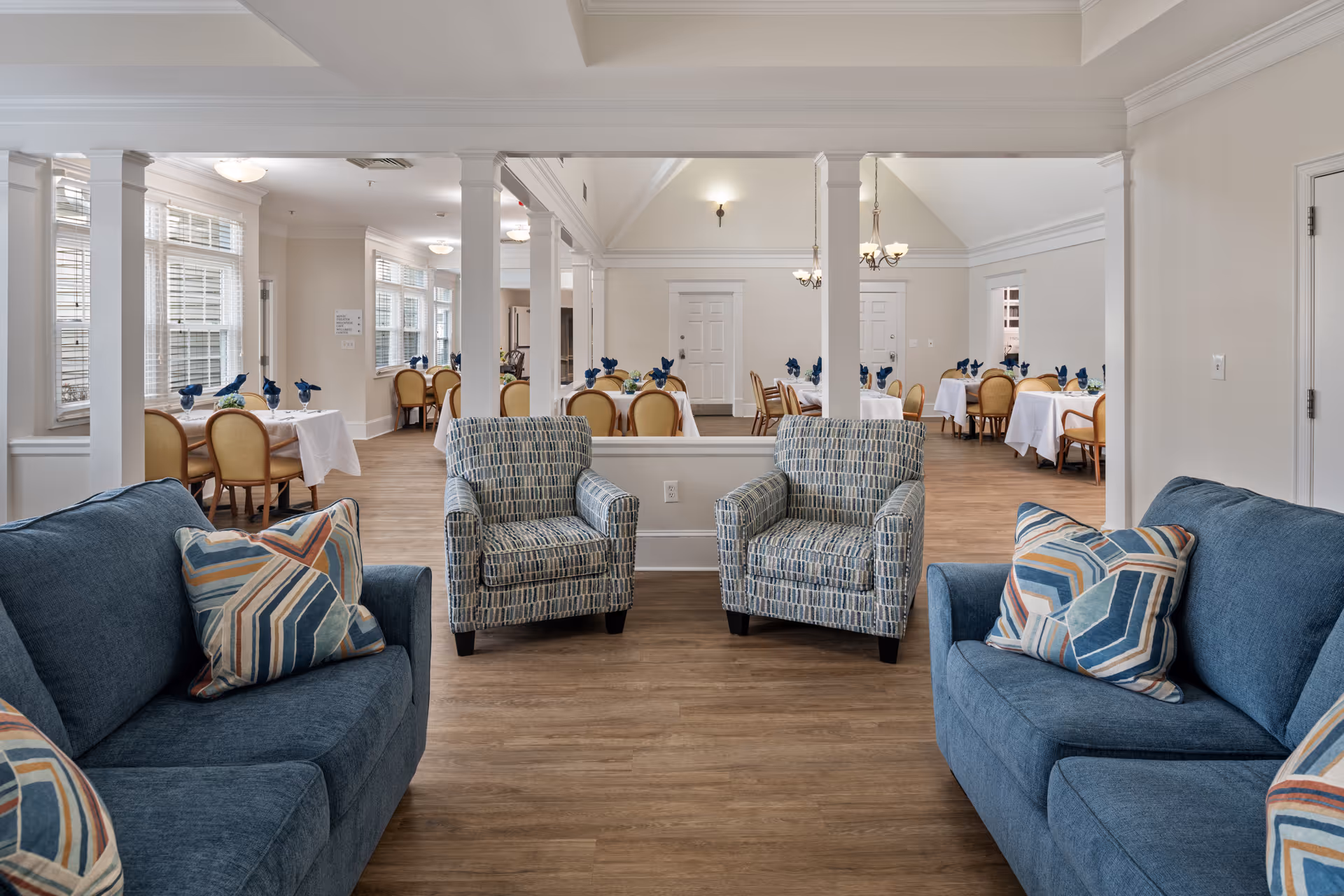 A bright and spacious common area in a senior living facility featuring two blue sofas with patterned pillows and two patterned armchairs arranged on a wood floor. Beyond the seating area, there is a dining room with multiple tables covered in white tablecloths, each set with blue napkins and chairs. The walls are painted light beige, and large windows allow natural light to fill the space.