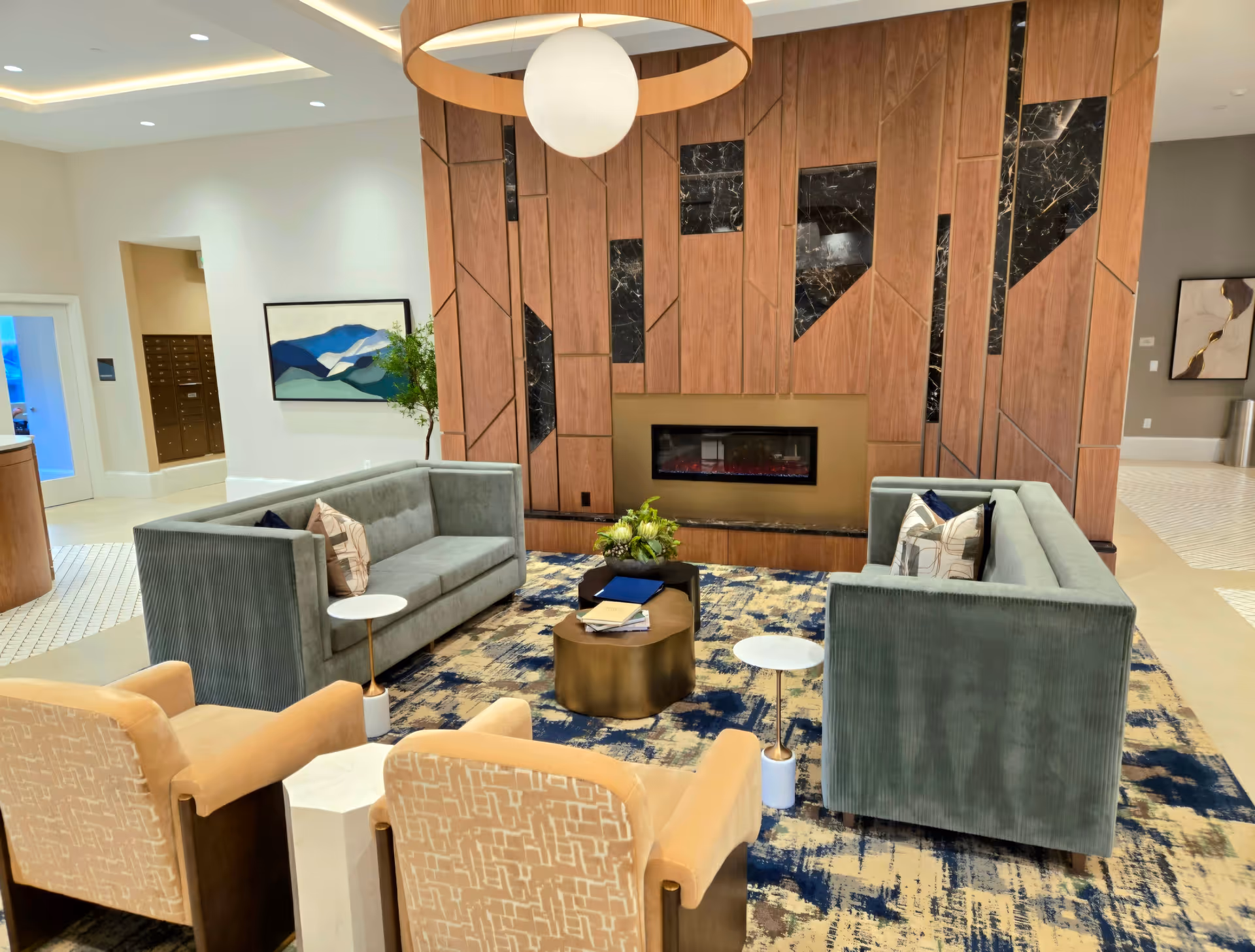 A modern and cozy lounge area featuring two green velvet sofas facing each other, two beige armchairs, a round gold coffee table with books and a plant, and a decorative fireplace set into a wooden and black marble accent wall. The room has a patterned carpet in shades of blue and beige, a large circular ceiling light, and artwork on the walls.
