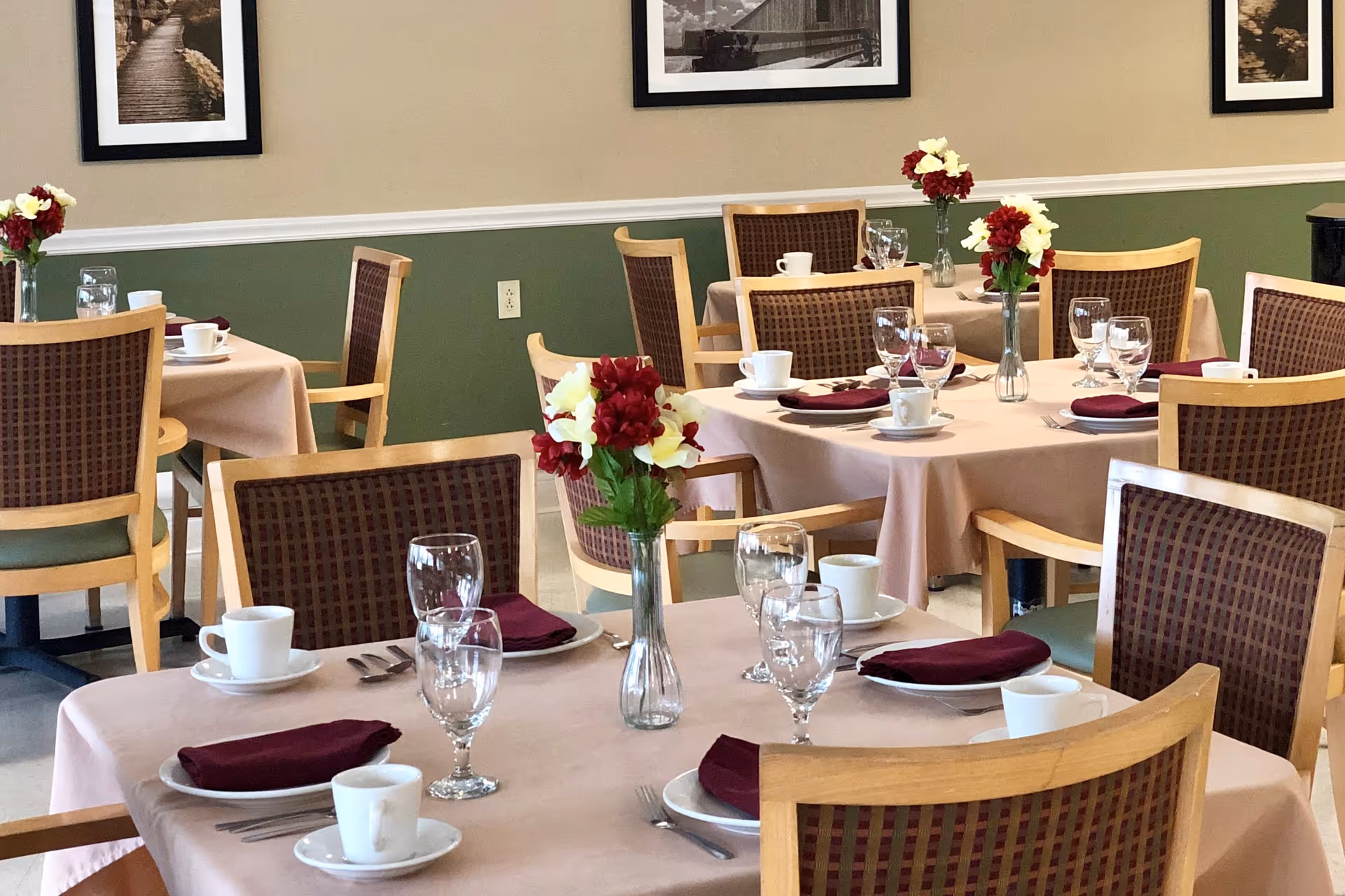 A dining room with several tables covered in light pink tablecloths, each set with white cups, plates, silverware, wine glasses, and burgundy napkins. Each table has a vase with red and white flowers. The walls are painted green with a beige upper section and decorated with framed black and white photographs.