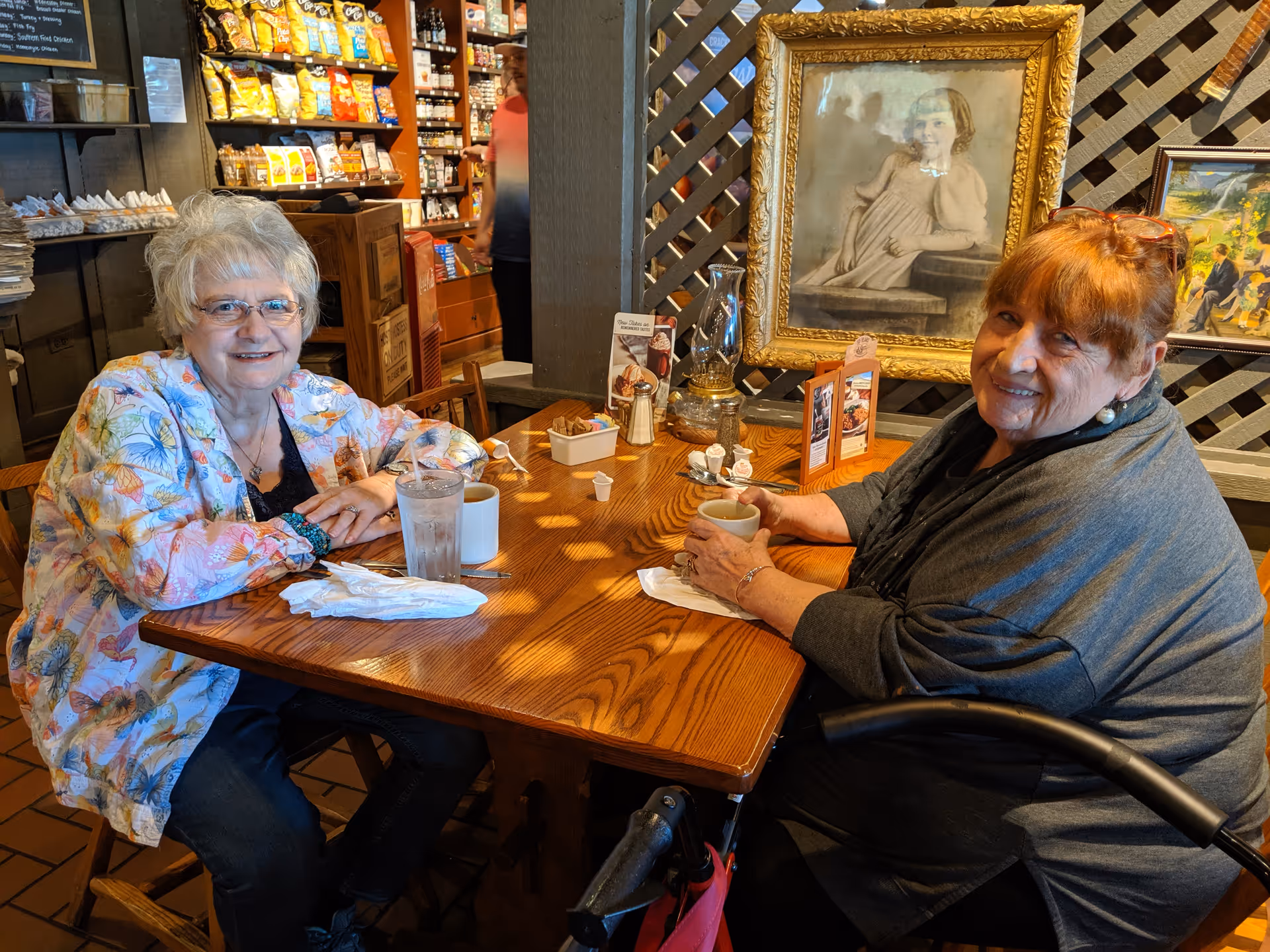 Two elderly women sitting at a wooden table in a cozy cafe or dining area. One woman with white hair and glasses wears a colorful floral jacket, while the other woman with reddish hair wears a dark shawl and holds a cup. Behind them are shelves stocked with snacks and other items, and a framed portrait of a young girl hangs on a lattice wall.