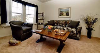 A cozy living room with a patterned sofa and a dark armchair arranged around a wooden coffee table. The coffee table holds a basket, a small floral arrangement, and some magazines. There is a large window with white curtains and dark drapes letting in natural light. A framed picture hangs on the wall above the sofa, and a tall vase with decorative branches is placed in the corner.