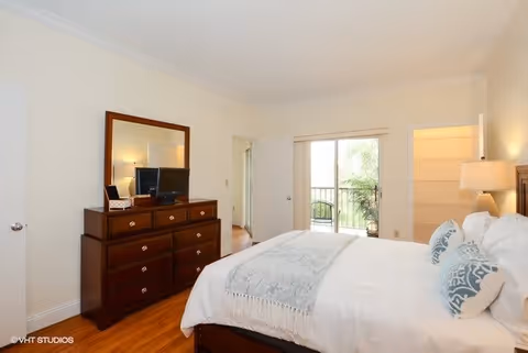 A bright bedroom with a large bed featuring white bedding and blue patterned pillows. There is a wooden dresser with a mirror and a small TV on top. The room has hardwood floors and a sliding glass door leading to a balcony with outdoor chairs and greenery visible outside.