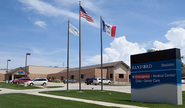 Exterior view of Sanford Health Sheldon Care Center building with a parking lot and several cars. Three flagpoles display the American flag, a white flag, and the Iowa state flag. A large sign in the foreground reads Sanford Sheldon Emergency Medical Center Clinic Senior Care.