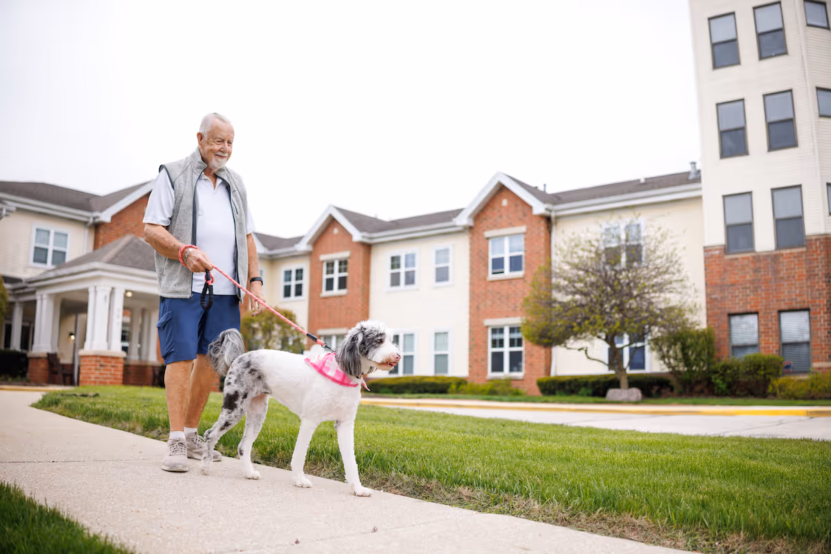 An elderly man walking a white and gray dog on a leash outside a multi-story senior living facility with brick and beige siding.
