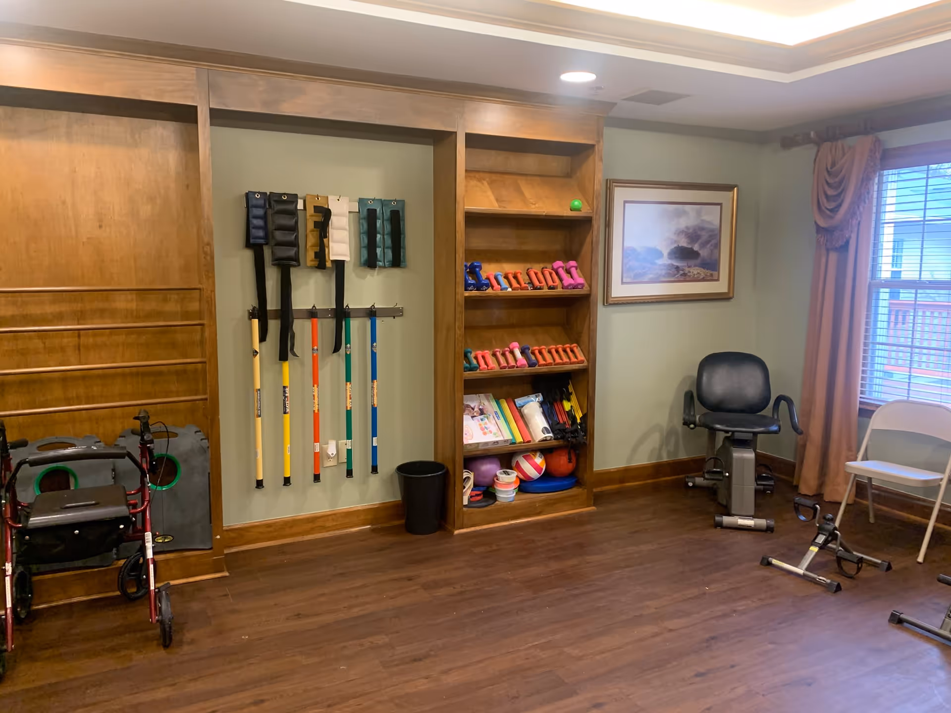 Small indoor exercise/therapy room with shelves of light dumbbells, colorful therapy poles, a recumbent exercise chair, a walker and folding chairs.