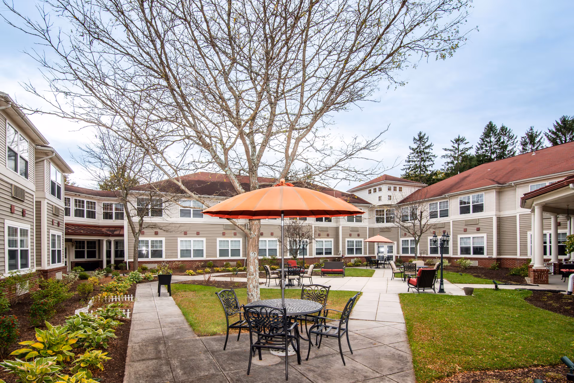 Outdoor courtyard area of a senior living facility with a round metal table and four chairs under an orange umbrella. The courtyard is surrounded by a two-story building with beige siding and red roofs. There are leafless trees, green grass, and paved walkways throughout the courtyard.