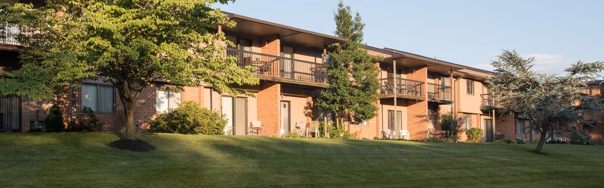 Exterior view of a two-story brick retirement community building with balconies and sliding glass doors, surrounded by green grass, trees, and shrubs under a clear sky.
