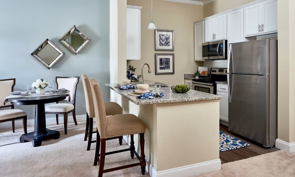 Open kitchen and dining area with a granite-topped island and bar stools, stainless steel appliances, and a small round dining table against a pale blue wall.