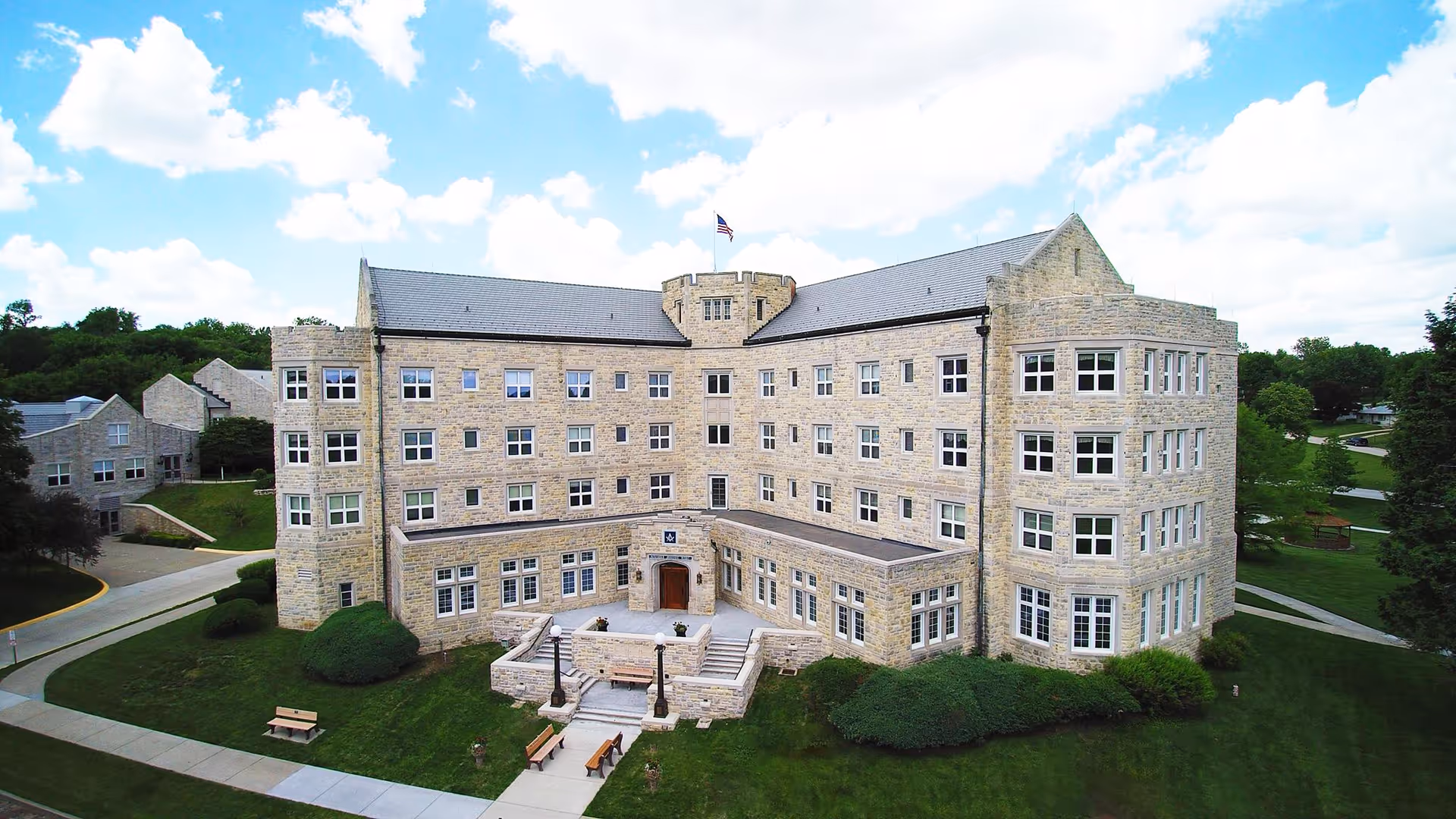 Exterior view of a large, multi-story stone building with many windows, a central entrance with steps, and an American flag flying on top. The building is surrounded by green lawns, trees, and paved walkways under a partly cloudy sky.