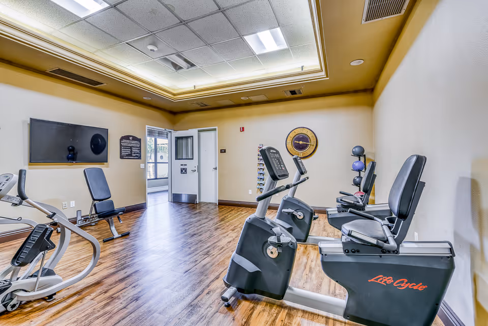 A small fitness room with exercise equipment including stationary bikes and a weight bench. The room has wood flooring, beige walls, a large clock on the wall, and a door leading to another area. The ceiling has recessed lighting with a decorative border.