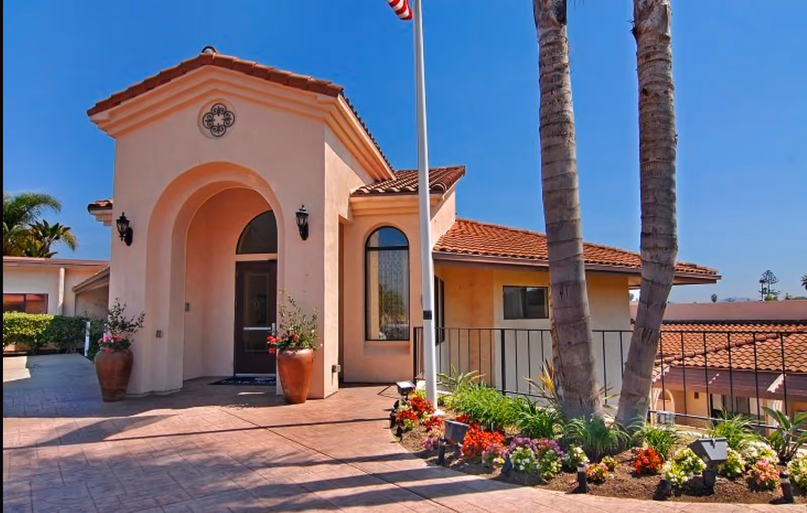 Exterior view of a senior living facility building with a peach-colored stucco facade, arched entrance, and red tile roof. There are two large potted plants with flowers flanking the entrance, a flagpole with an American flag, two tall palm trees, and a landscaped flower bed with colorful flowers in front of the building under a clear blue sky.