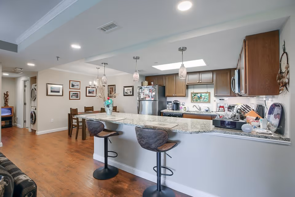 Interior view of a kitchen and dining area in a senior living facility. The kitchen features wooden cabinets, a stainless steel refrigerator, a microwave, and various kitchen appliances on the countertops. A granite countertop bar with two modern bar stools separates the kitchen from the living space. The dining area has a wooden table with chairs and framed pictures on the wall. Hardwood floors and recessed lighting complete the space.