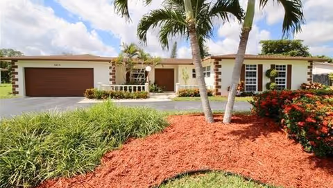 Single-story residential building with a brown garage door, white exterior walls with brown trim, and a landscaped front yard featuring palm trees, green shrubs, and red mulch under a partly cloudy sky.