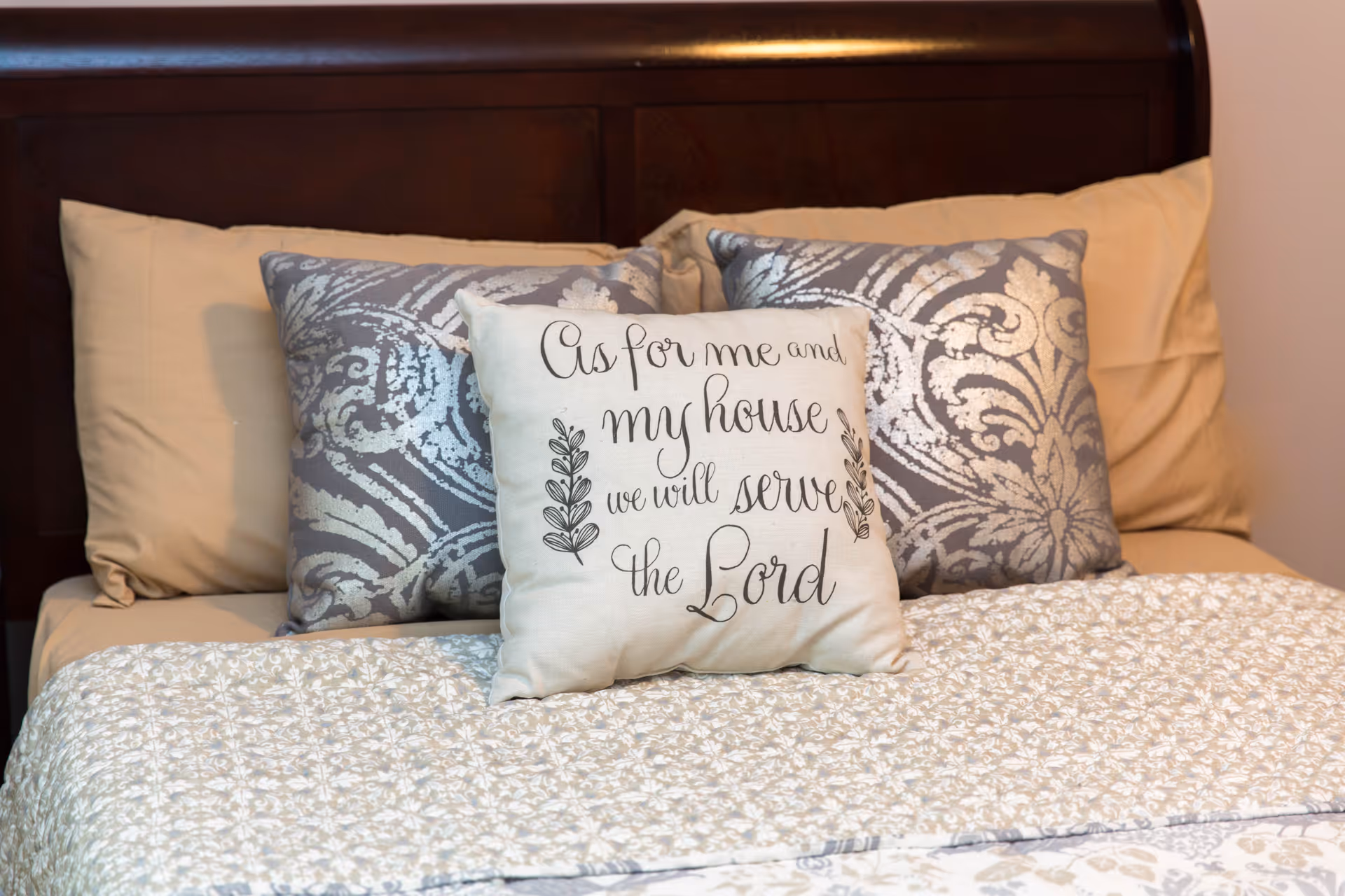 A neatly made bed with beige sheets and a quilted bedspread featuring a subtle floral pattern. There are four pillows arranged against a dark wooden headboard: two beige pillows at the back, two decorative pillows with a silver and gray damask pattern in front, and a smaller white pillow in the center with the text 'As for me and my house we will serve the Lord' written in cursive.