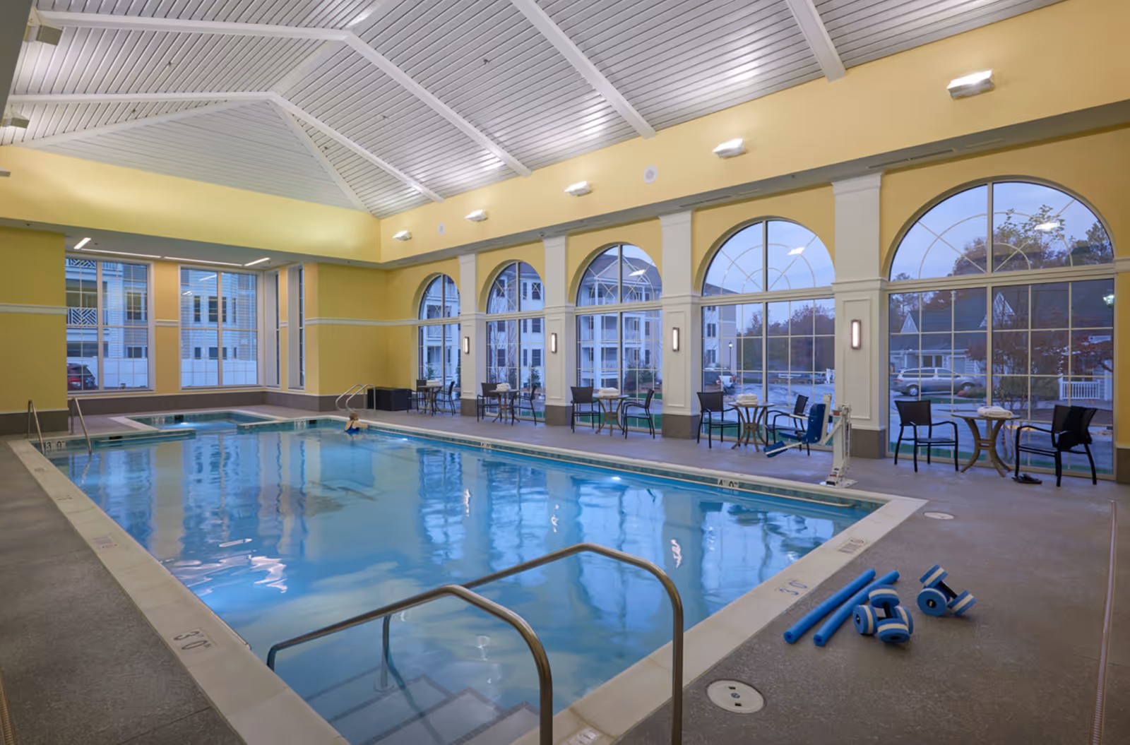 Indoor swimming pool area with a person in the water, surrounded by large arched windows showing an outside view of buildings and trees. Poolside has chairs and small tables with towels, and pool exercise equipment is placed on the floor near the pool edge. The ceiling is high with white beams and yellow walls.