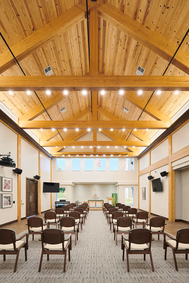 Interior view of a chapel or meeting room with wooden vaulted ceiling and exposed beams, rows of wooden chairs with cushions arranged facing an altar at the front, large windows above the altar letting in natural light, and two wall-mounted televisions on either side.