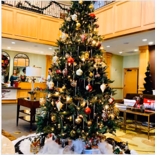 A large decorated Christmas tree with ornaments and lights stands in the center of a spacious, warmly lit room. The room features wooden cabinetry, a mirror, a table with a lamp, and seating areas with cushions. Holiday decorations and wrapped gifts are placed around the base of the tree.