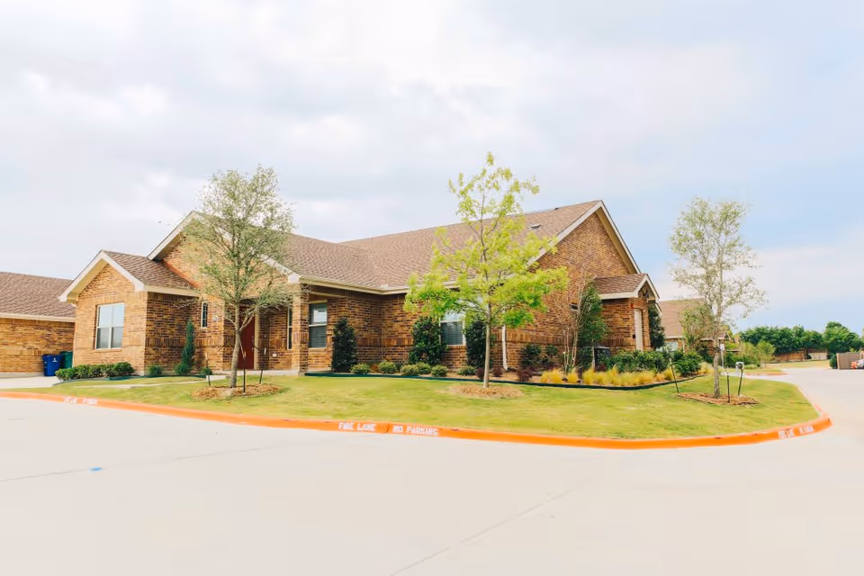 Exterior view of a single-story brick building with a sloped roof, surrounded by small trees and landscaped grass areas. The building is located on a paved street with a curb painted orange marked as a fire lane with no parking.