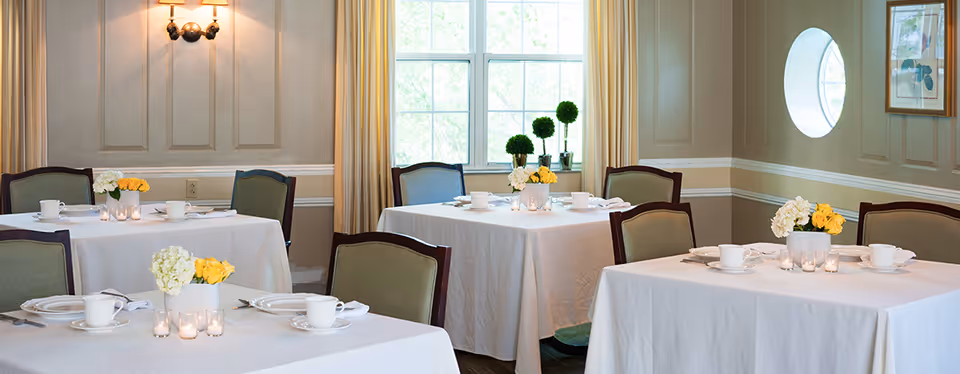 A bright dining room with several tables covered in white tablecloths, each set with white cups, plates, napkins, and small floral centerpieces. The room has beige walls with white trim, a window with beige curtains, a round window, and a wall sconce light fixture.